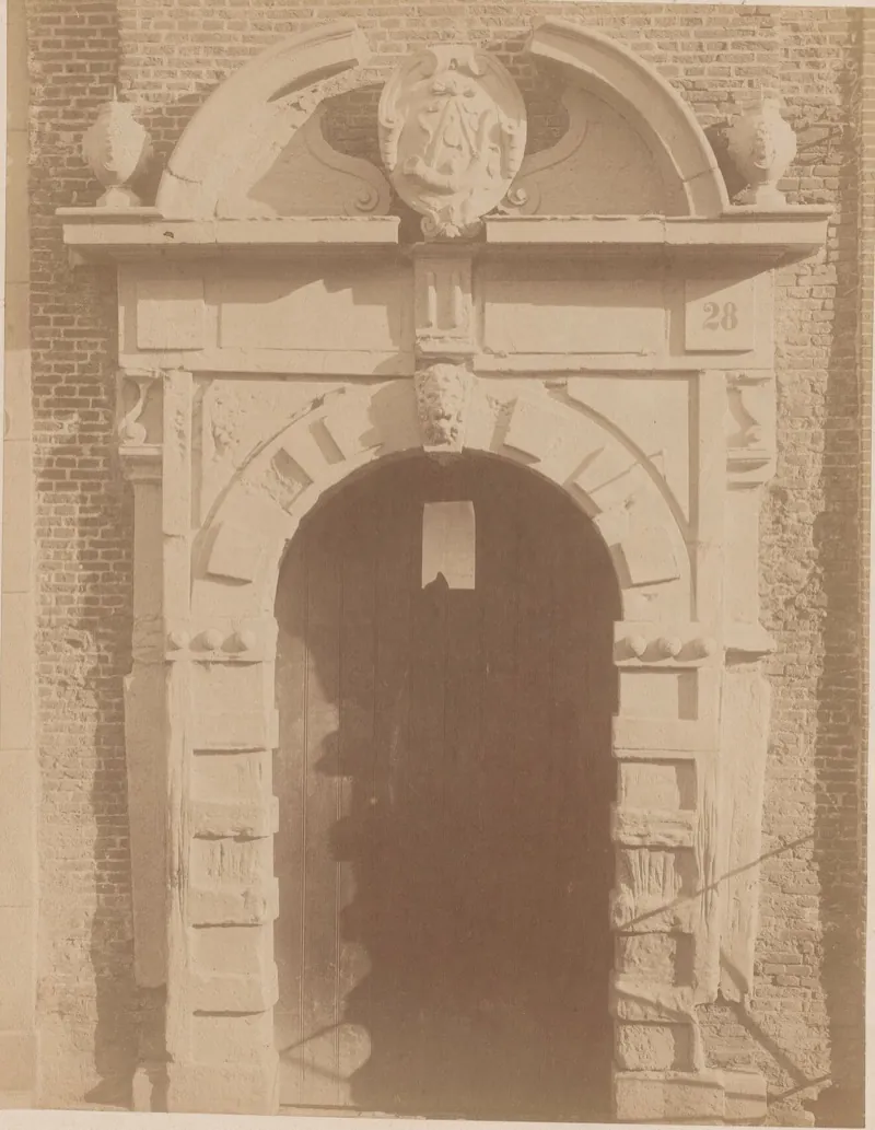 Un haut portail de pierre à arc en plein cintre, surmonté d’armoiries sculptées dans un fronton brisé, deux vases de pierre sur les côtés et le numéro 28 en haut à droite, dans un mur de briques.
