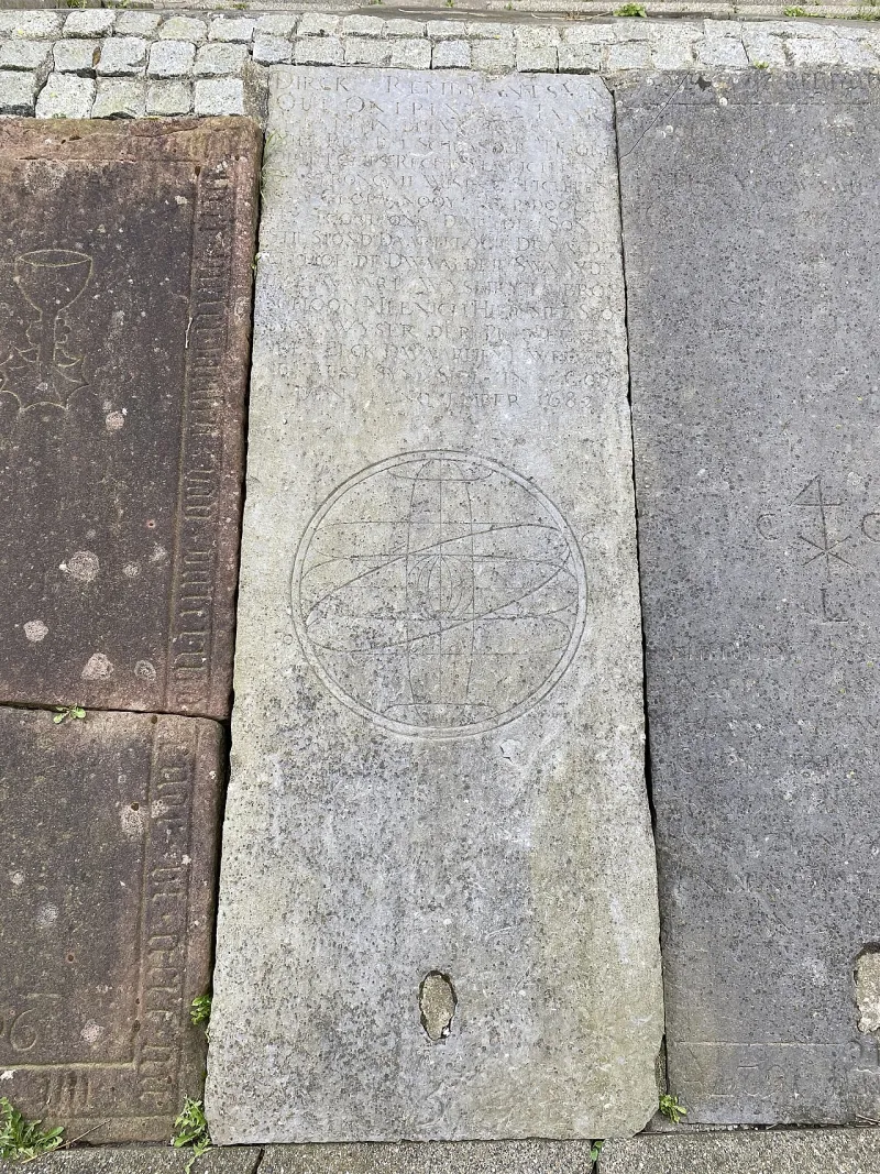 Eine graue Grabplatte zwischen anderen Platten in einem gepflasterten Weg. Oben eine verwitterte Inschrift in Großbuchstaben, darunter ein großer eingemeißelter Kreis mit einer von Ringen durchzogenen Erdkugel.