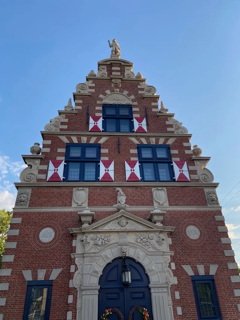 La façade du Zwaanendael Museum à Lewes vue d’en bas sur un ciel bleu : pignon à gradins en briques rouges, bandeaux de pierre blanche, volets rouges et blancs, portail sculpté et, au sommet, la statue de De Vries.