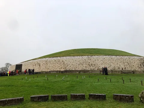 Le côté sud-est de Newgrange : un tertre bas, rond et recouvert d’herbe, avec une façade de quartz blanc et de galets de granite, et à gauche l’entrée sombre avec un escalier et des visiteurs.