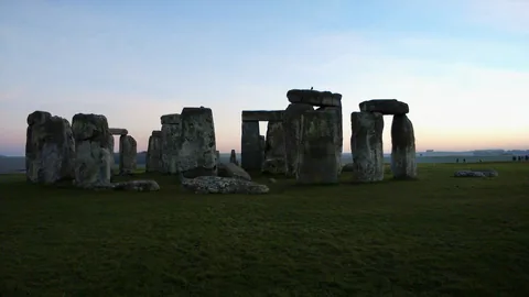 Le cercle de pierres de Stonehenge dans une douce lumière du soir : pierres dressées sombres surmontées de linteaux sur un ciel pâle, de l’herbe au premier plan.